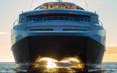 Bow of mega catamaran Elite with dark hull & white upper decks, sitting in calm water with sunset light shining underneath.