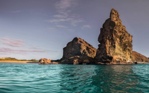 Turquoise water beside white-sand beach & 2 large rock outcrops reaching to the sky, seen on the Elite Galapagos cruise.