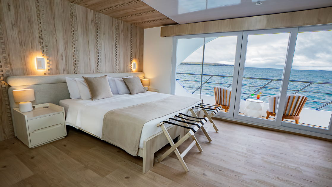 Double bed with white, beige & teal bedding in room with white & wood accents, wooden desk & shelves & glass door onto private balcony.