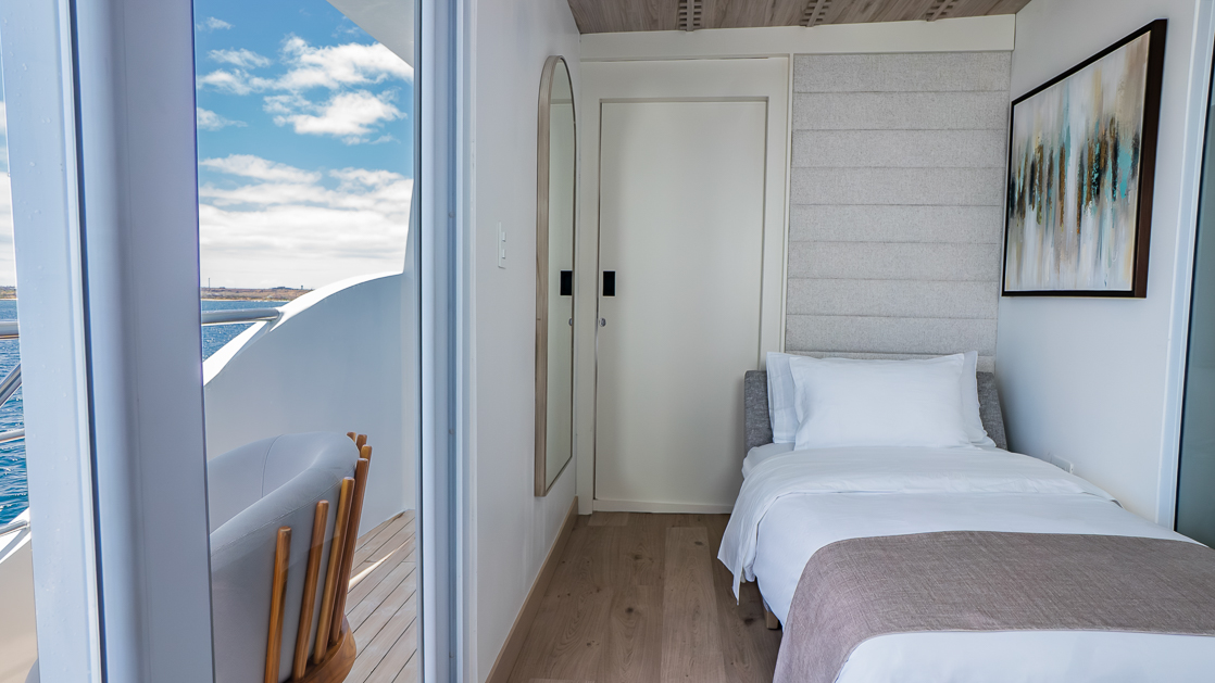 Twin bed with white, beige bedding in slender room with white & wood accents aboard Endemic cruise ship in the Galapagos.