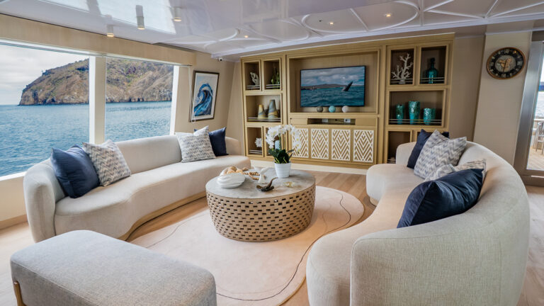 Double bed with white, beige & teal bedding in room with white & wood accents, wooden desk & shelves & glass door onto private balcony.