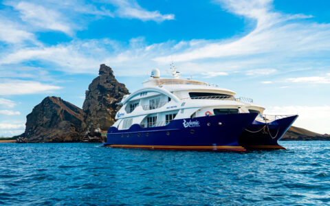 Endemic Galapagos catamaran with blue hull & 2 white upper decks sits in calm sea beside jagged dark rocks under sunny skies.