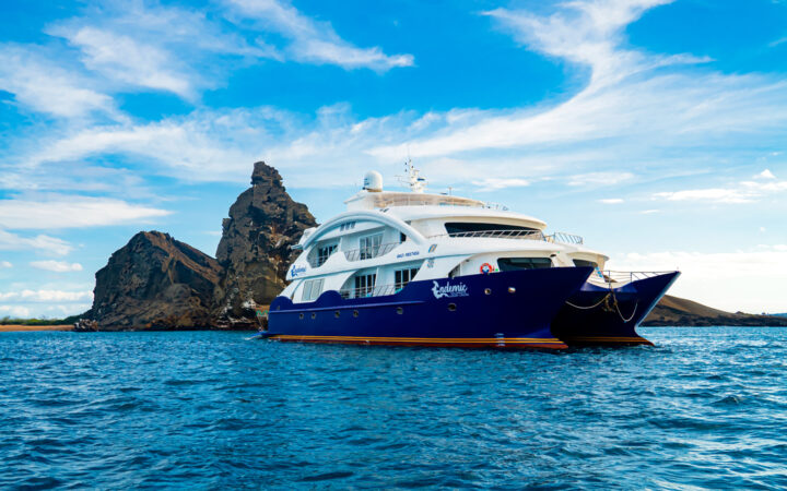 Endemic Galapagos catamaran with blue hull & 2 white upper decks sits in calm sea beside jagged dark rocks under sunny skies.