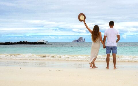 Man & woman in light-colored clothes stand on white-sand beach waving to white yacht on a Grand Majestic cruise in Galapagos.