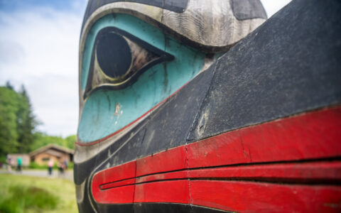 Close up of a carved wooden totem pole with bird-like face, painted teal, black & red in Alaska.