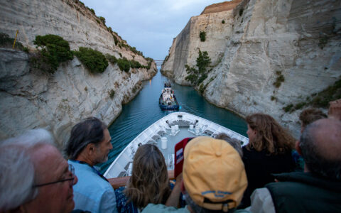 Travelers watch from the upper deck of a white small ship as a tug boat pulls them through the Corinth Canal's beige walls.