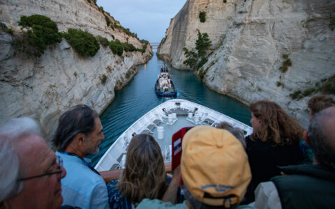 Travelers watch from the upper deck of a white small ship as a tug boat pulls them through the Corinth Canal's beige walls.