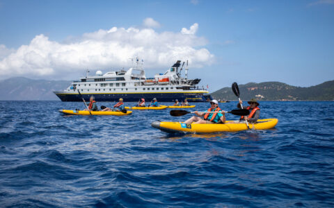 Travelers paddle inflatable yellow tandem kayaks near a National Geographic small ship in choppy blue sea on a sunny day.