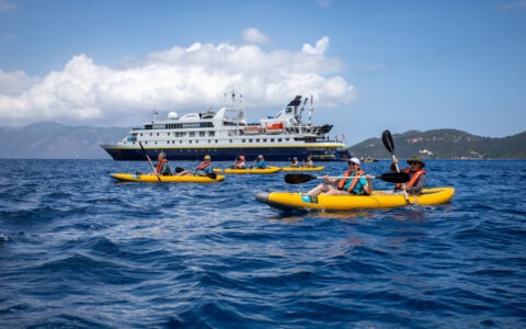 Travelers paddle inflatable yellow tandem kayaks near a National Geographic small ship in choppy blue sea on a sunny day.