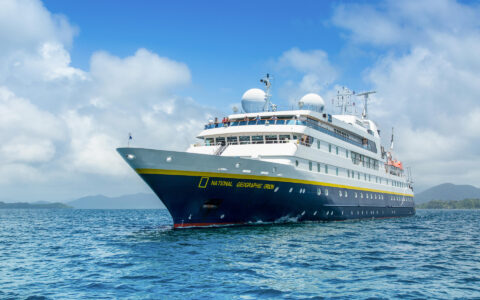 National Geographic Orion small ship with dark blue hull & white upper decks sits at sea in sunshine as guests wave from bow.