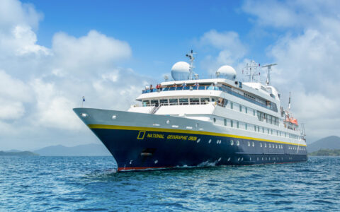 National Geographic Orion small ship with dark blue hull & white upper decks sits at sea in sunshine as guests wave from bow.