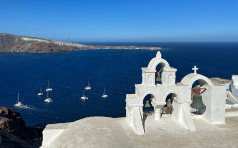 Whitewashed bell towers look out over deep blue Aegean Sea with small white sailboats & volcanic land behind, in Santorini.