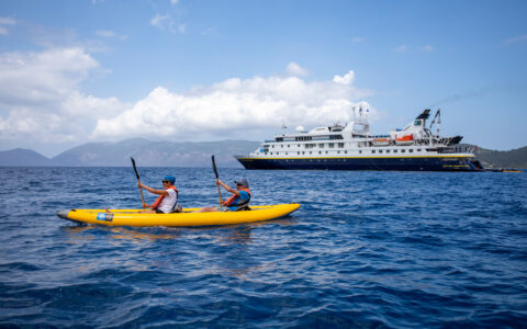 A pair of tandem kayakers in an inflatable yellow boat paddle calm blue sea beside the National Geographic Orion small ship.