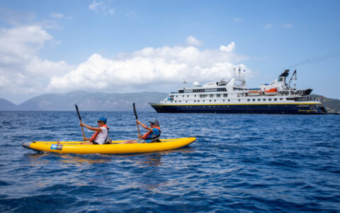 A pair of tandem kayakers in an inflatable yellow boat paddle calm blue sea beside the National Geographic Orion small ship.