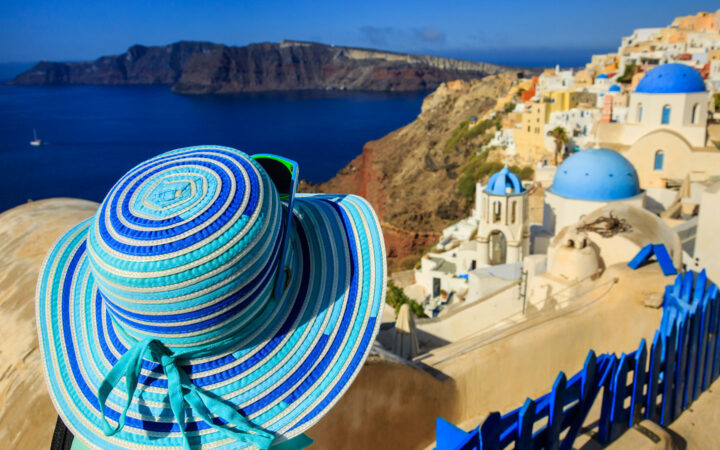 Traveler in wide-brimmed blue & turquoise sun hat looks out over Santorini mountaintop town with blue domes & beige buildings.
