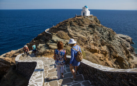 Greece travelers traverse a stone walkway along the spine of a cliff, towards a blue-domed, whitewashed church by the sea.