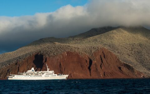 The all white Galapagos small ship the Legend floats in the dark blue ocean against a deep red hillside of Rabida island.