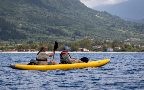 Tandem kayakers weiring beige paddle an inflatable yellow boat along lush green shores on a hot summer day in Greece.