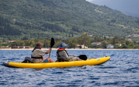 Tandem kayakers weiring beige paddle an inflatable yellow boat along lush green shores on a hot summer day in Greece.