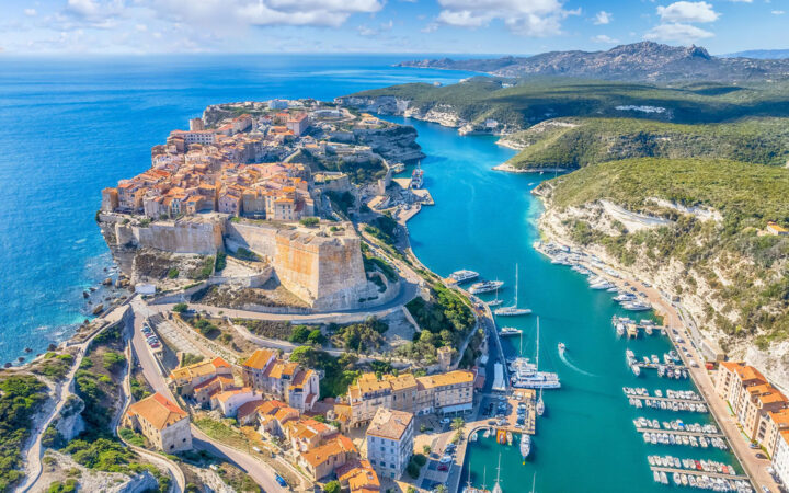 Aerial view of a Mediterranean port town showing boats docked at a castle and red roof homes on an island