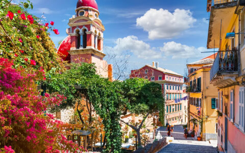 Greek street with colorful buildings and bright blooming greenery around the cobblestone path.