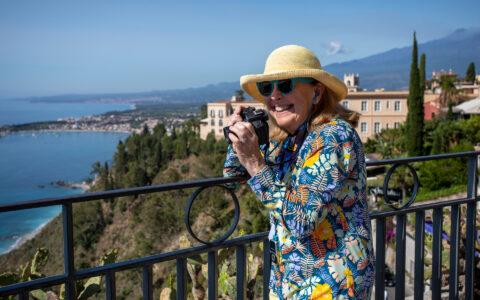 Woman in multi-color floral shirt holds camera to photograph Taormina from an overlook with a black wrought-iron fence.