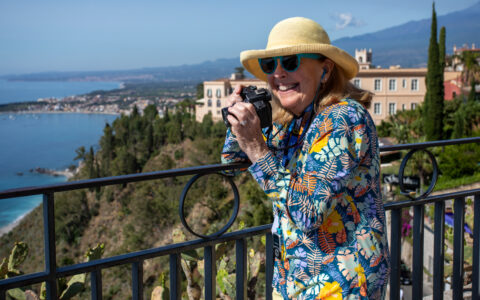 Woman in multi-color floral shirt holds camera to photograph Taormina from an overlook with a black wrought-iron fence.