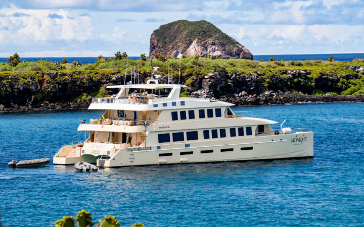 Aerial view of Monique small ship cruising the calm waters of the Galapagos with vegetation-rich land mass in the background.