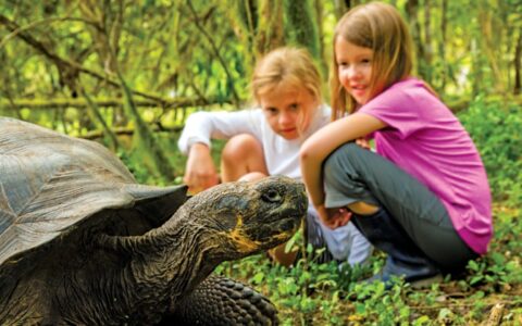 2 young girls crouch down beside a giant tortoise among bright green grass and moss during the Wild Galapagos Escape Cruise