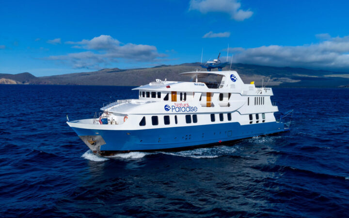 Exterior port side of Natural Paradise Galapagos small ship cruising with blue hull & 3 white upper decks.