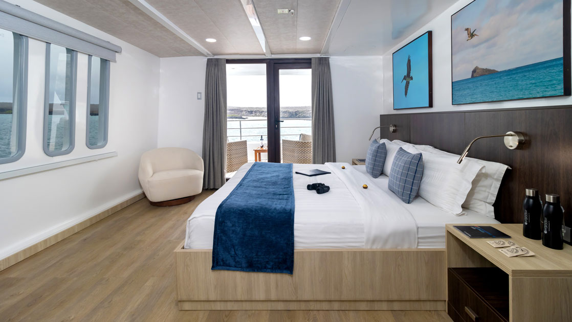 Upper Deck Suite with king bed in blue & white linens, framed Galapagos photos & private balcony on Natural Paradise yacht.