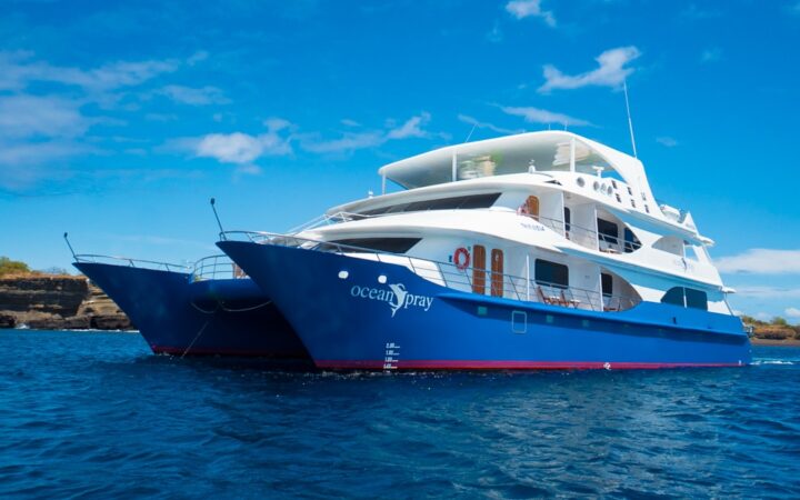 Small catamaran Ocean Spray with blue hull & white upper decks sits in calm water on a sunny day in Galapagos.