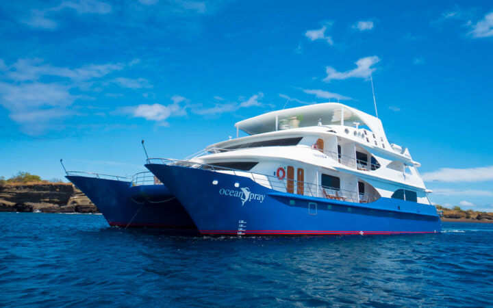 Small catamaran Ocean Spray with blue hull & white upper decks sits in calm water on a sunny day in Galapagos.