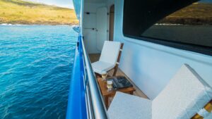 Narrow private balcony with 2 padded wooden chairs & wood coffee table on a ship sitting in calm water on a sunny day.