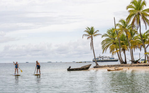 Man & woman paddle stand-up paddlebaords by shore of tiny island with white-sand beach & palm trees as ship sits nearby in Panama.