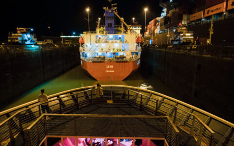 Guest stands on bow of small ship Nat Geo Quest as she transits the Panama Canal behind a cargo ship with red hull at night.