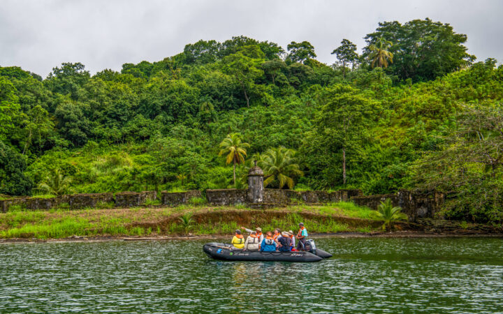 Zodiac boat with Panama canal travelers motors past aging historic fort walls backed by jungle as a guide points.