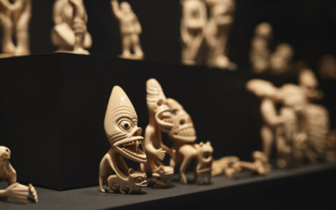 Small beige carved characters with peaked heads, large eyes & wide mouths sit on display in a South Greenland museum.