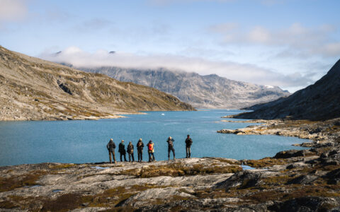 Small group of hikers stand atop a rocky outcrop overlooking a fjord with deep blue water flanked by rocky mountains.