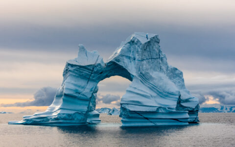 Large iceberg with natural arch & striations of blue & black rises out of calm sea, seen on a South Greenland cruise.