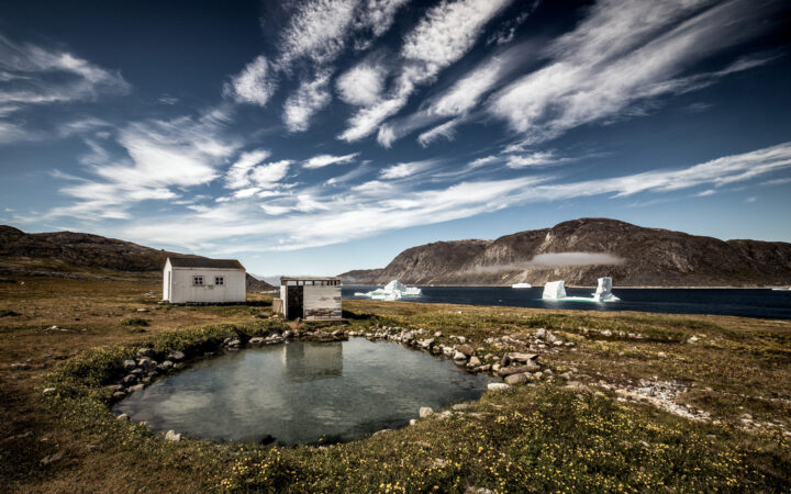 Natural hot spring with weathered small building beside fjord with large white icebergs & mountains behind in Greenland.