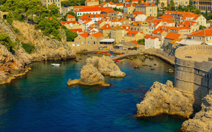 Blue-green Aegean Sea touches Dubrovnik Croatia's cove with kayakers & sun seekers below terracotta roofs.
