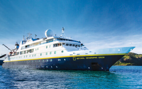 National Geographic Orion small ship with dark blue hull, yellow accents & white upper decks sits in turquoise sea in the sun.