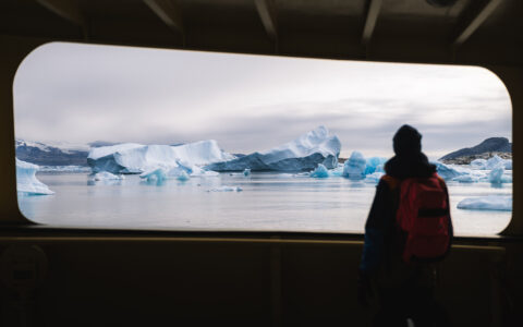 West Greenland traveler on the Disko Bay to Uummannaq cruise stands on deck of ship & views blue icebergs.