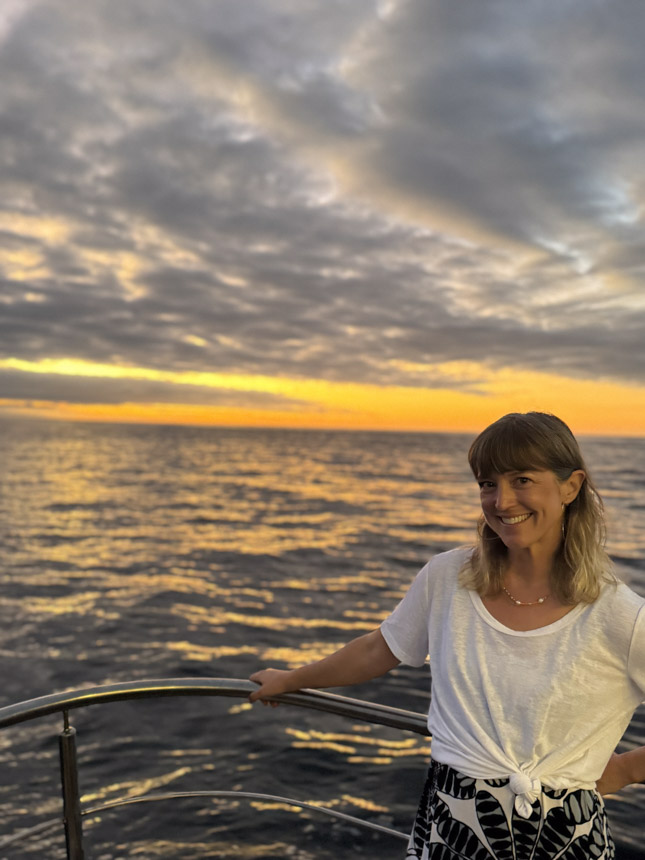 Woman in white t-shirt smiles standing on deck of a small ship at sea during an orange & yellow sunset.