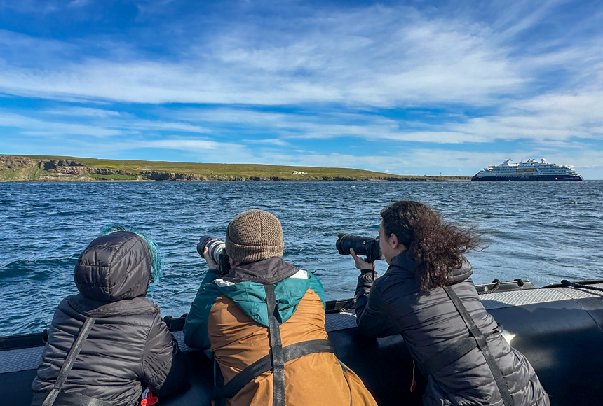 Three people looking out from small zodiac over the waters of Iceland with a small nat geo ship off in the distance on a sunny day