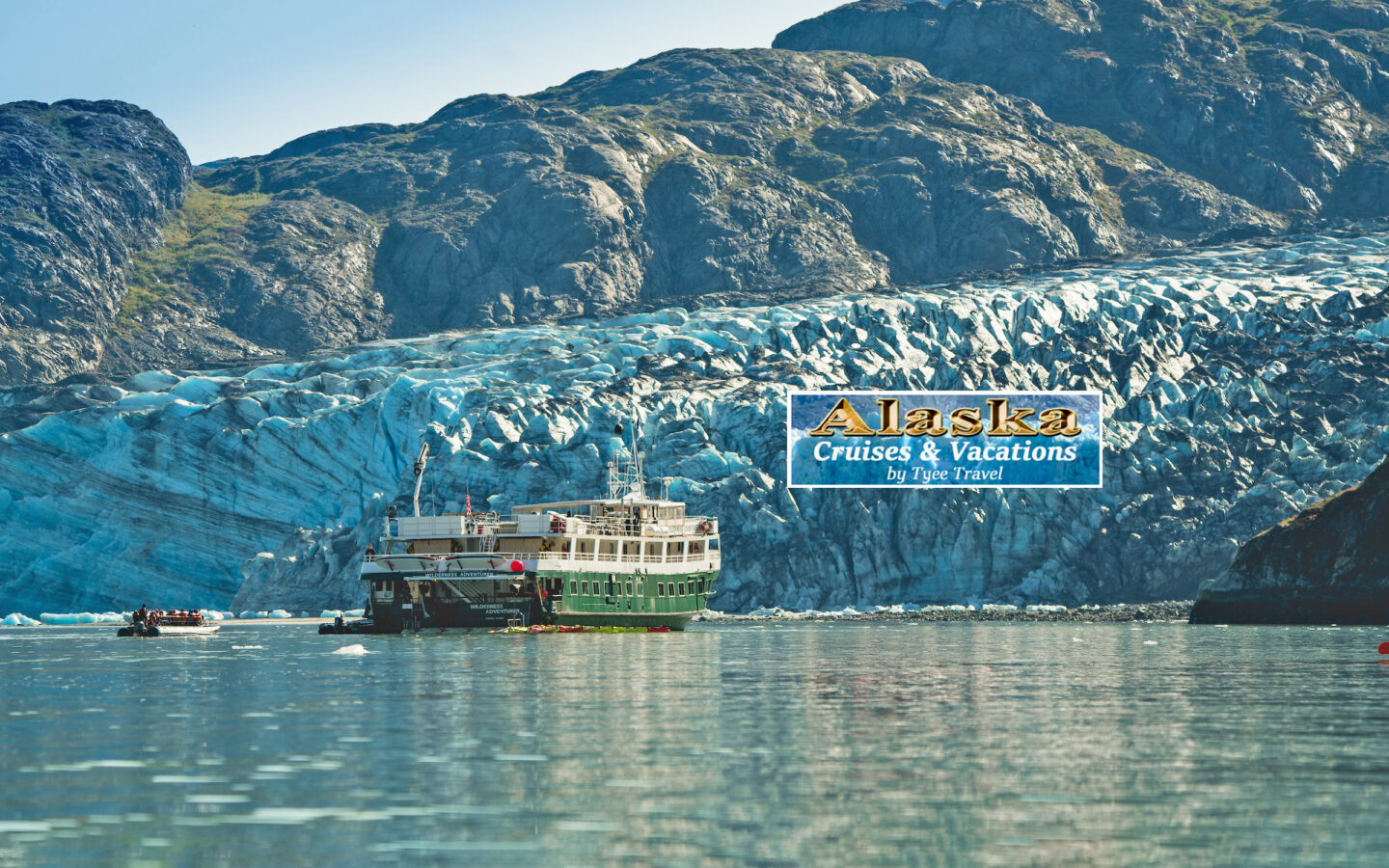 Green and white small ship Wilderness Adventurer anchored in front of teal glacier in Alaska on sunny day.