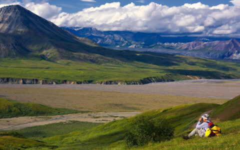 Alaska traveler sits on a grassy mountainside looking at mountains across a big river basin on the Alaska Coast to Denali Journey.