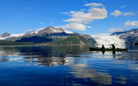 Tandem kayakers paddle glassy water backed by a white glacier & green hills under a blue sky on the Alaska Coast to Denali Journey.