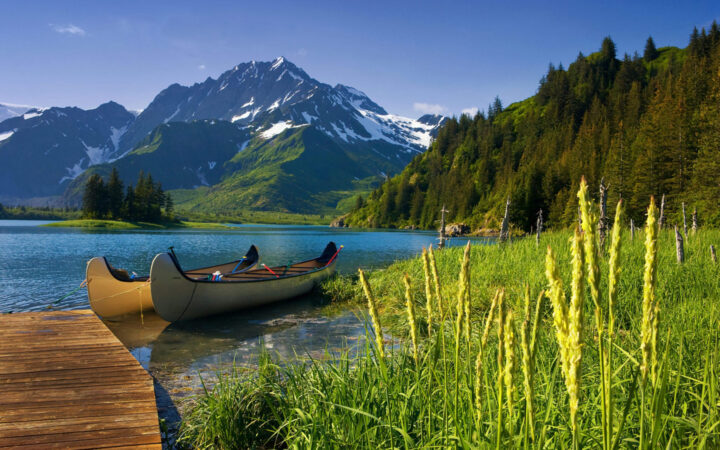 In Alaska. two canoes are tied up to a dock that extends into a blue lake surrounded by jagged snow capped mountains and dense forested hillside.